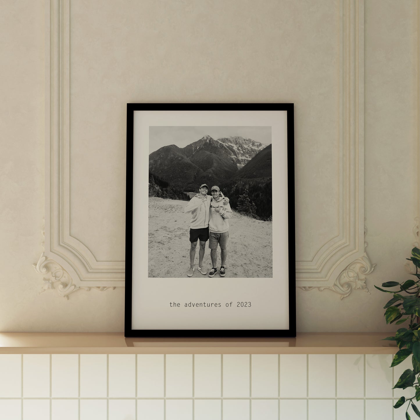 Custom Monochrome Photo Print featuring personalised photo of 2 men with arms around each other. Features custom text saying "the adventures of 2023". Print is framed in a black frame and displayed on a ledge in an expensive looking room.