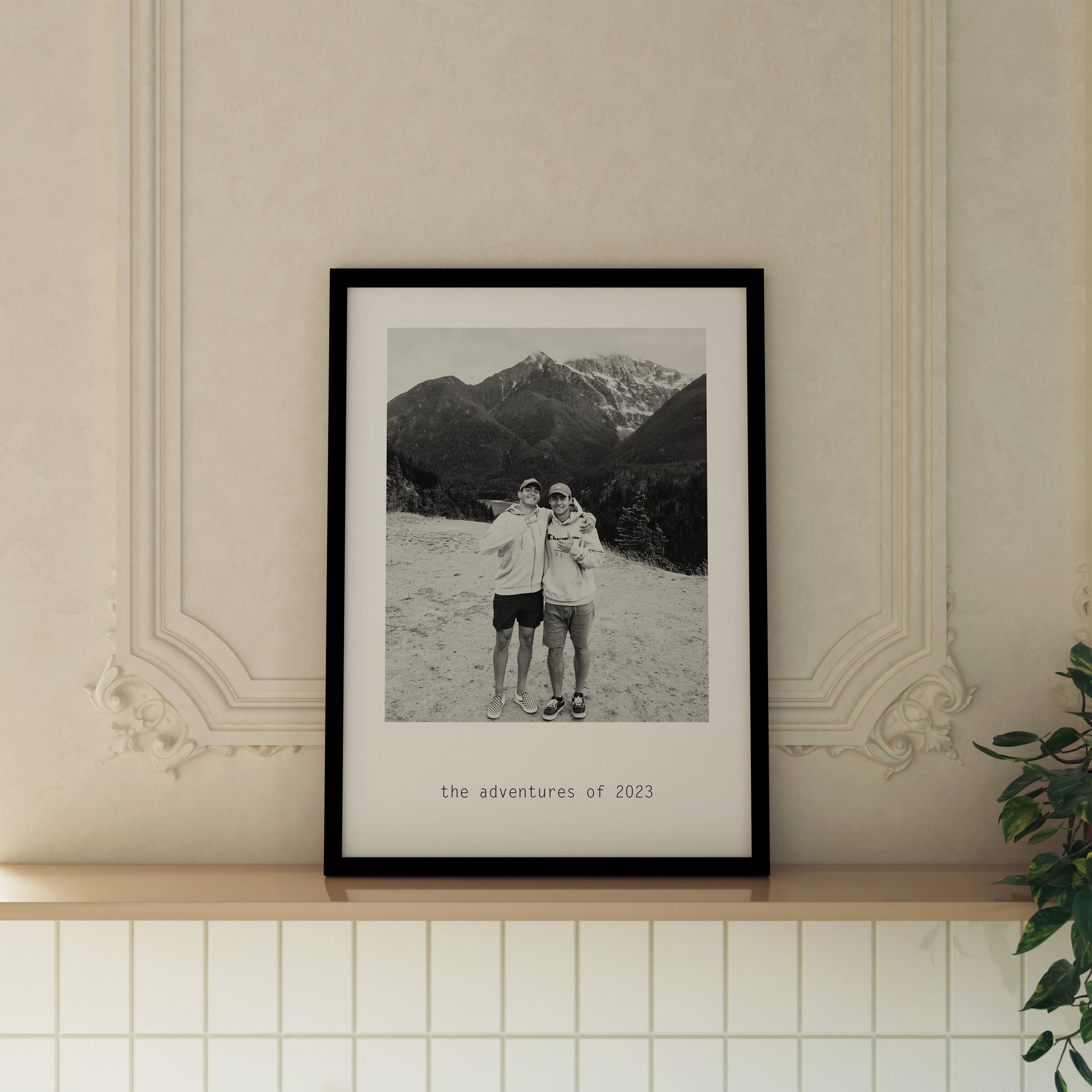 Custom Monochrome Photo Print featuring personalised photo of 2 men with arms around each other. Features custom text saying "the adventures of 2023". Print is framed in a black frame and displayed on a ledge in an expensive looking room.