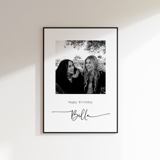Print featuring black and white photo sent by customer. Photo features 2 girls laughing. Custom text is added below of your own choosing, here featuring "happy birthday" in small text and "Bella" in large script text below. Print is framed in a thin black frame