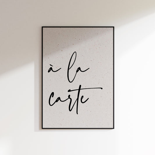A print on textured paper with black text that reads 'à la carte' in a handwriting style typeface. A culinary print for the kitchen. Print is in a black frame on a white wall background.