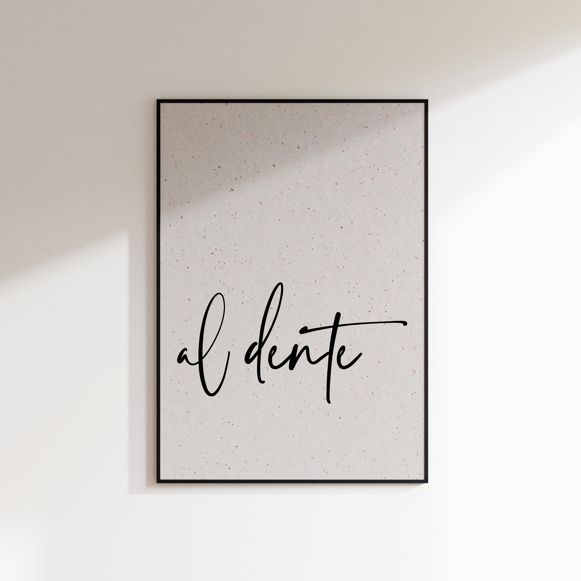 A print on textured paper with black text that reads 'al dente' in a handwriting style typeface. A culinary print for the kitchen. Print is in a black frame on a white wall background.