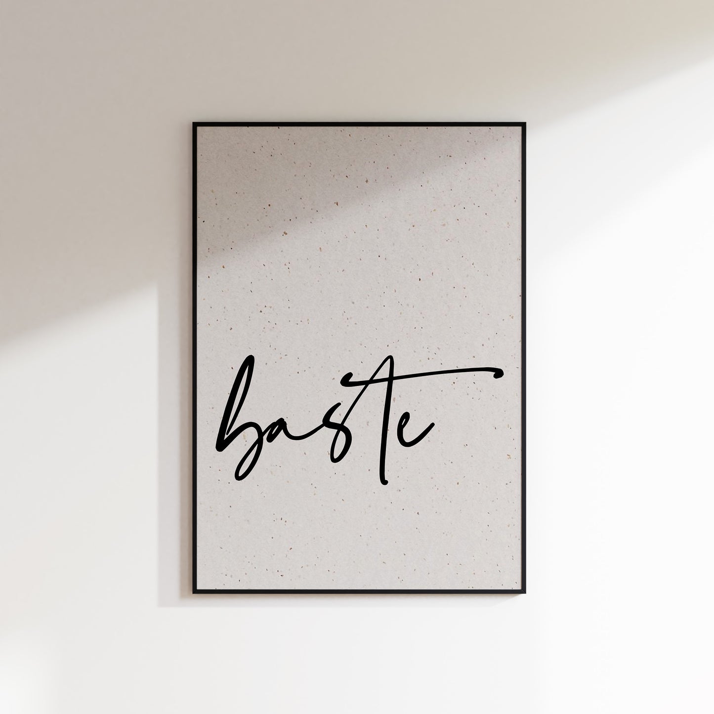 A print on textured paper with black text that reads 'baste' in a handwriting style typeface. A culinary print for the kitchen. Print is in a black frame on a white wall background.