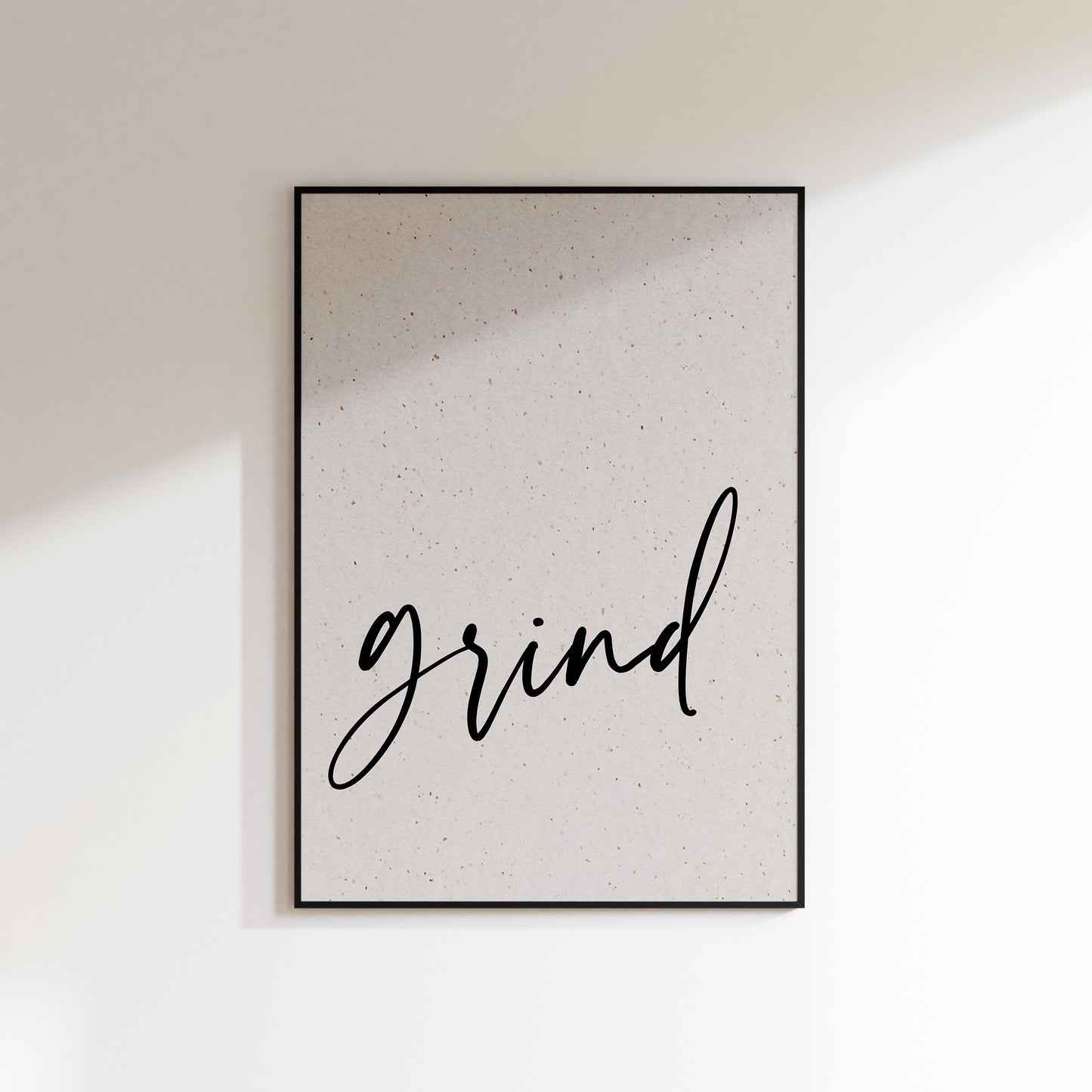 A print on textured paper with black text that reads 'grind' in a handwriting style typeface. A culinary print for the kitchen. Print is in a black frame on a white wall background.