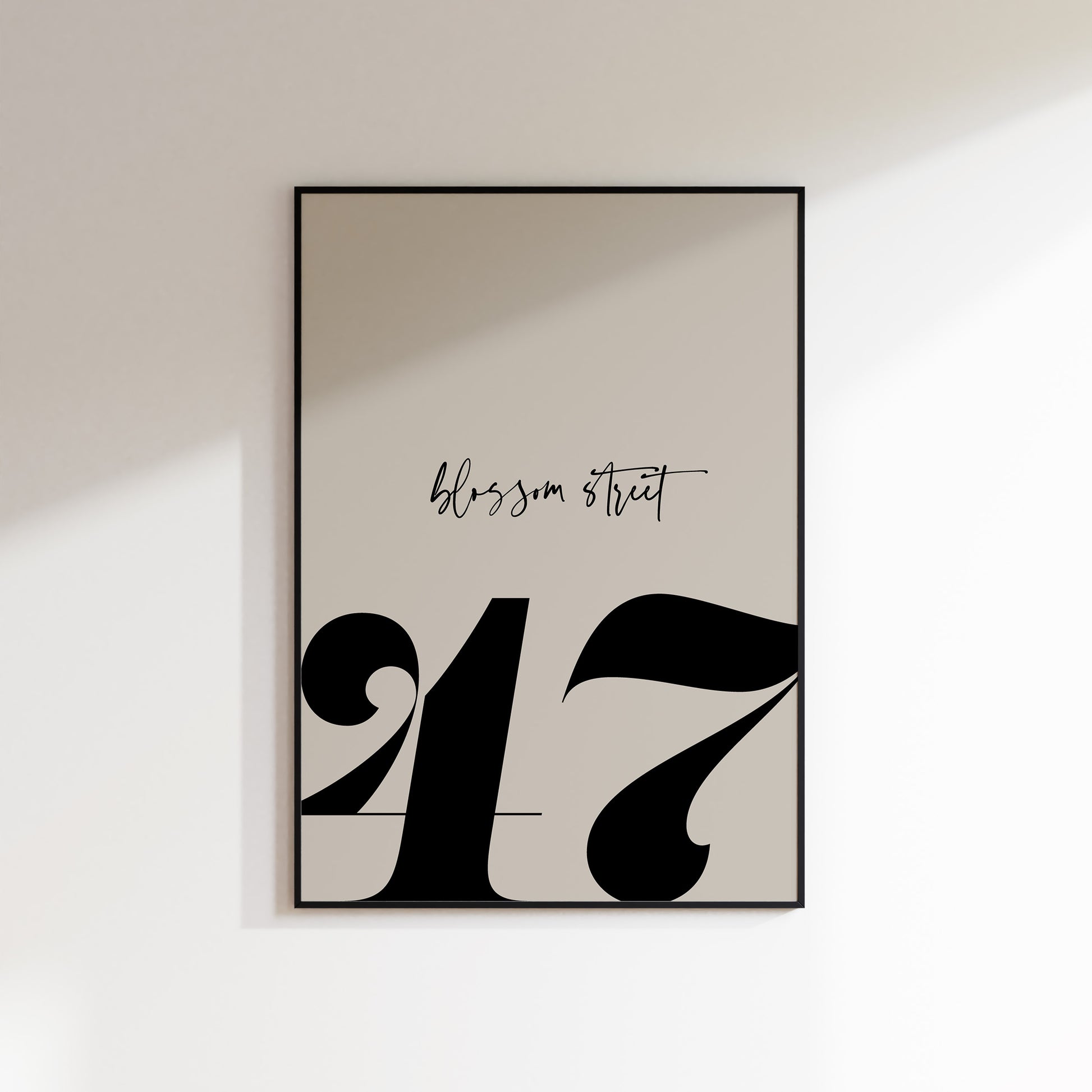 A house number print for the home, ideal for hallways, living rooms and bedrooms. This is a neutral beige coloured print with a slate coloured house number in a bold serif typeface, and the street name above in a handwriting style typeface. Print is in a black frame on a white wall background.
