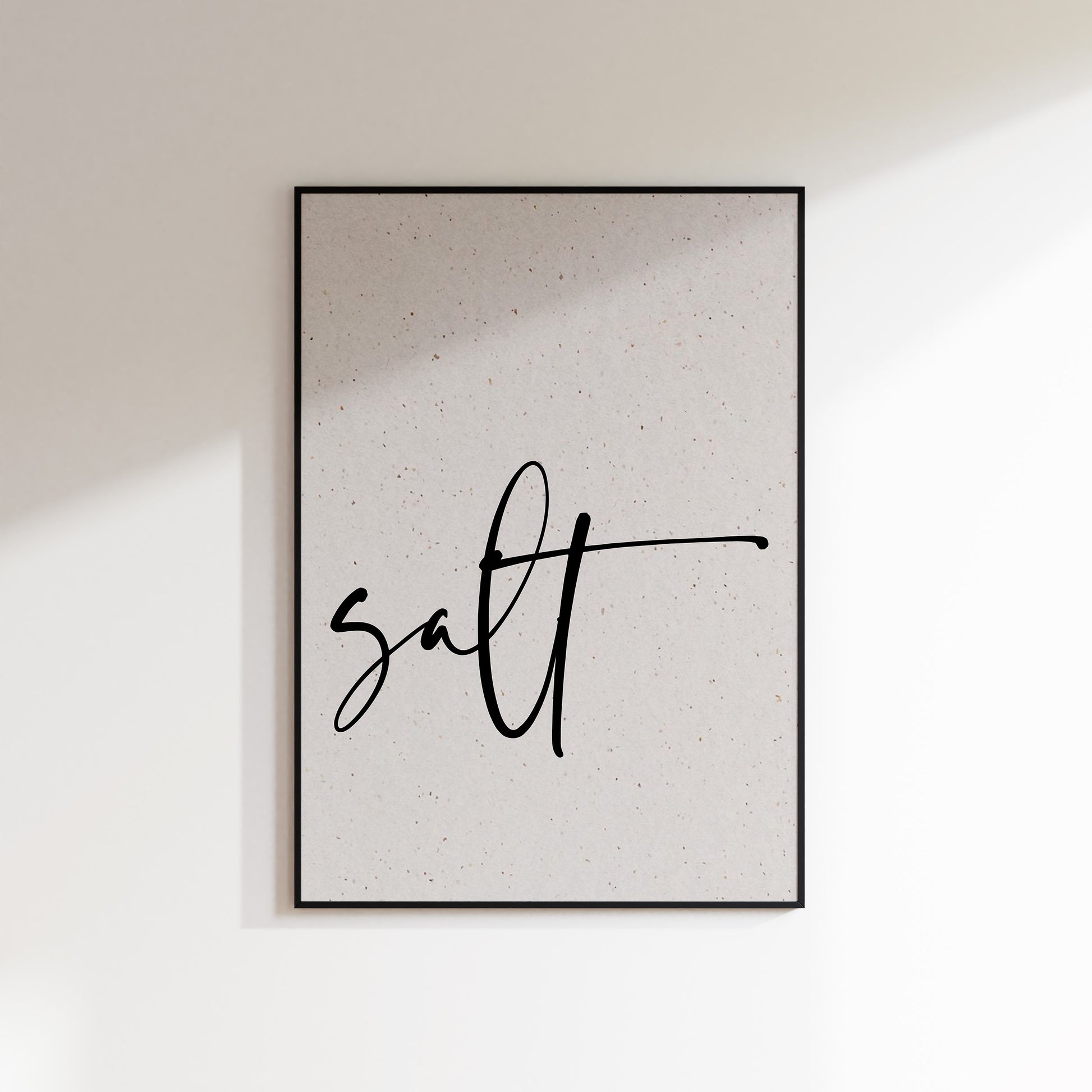 A print on textured paper with black text that reads 'salt' in a handwriting style typeface. A culinary print for the kitchen. Print is in a black frame on a white wall background.