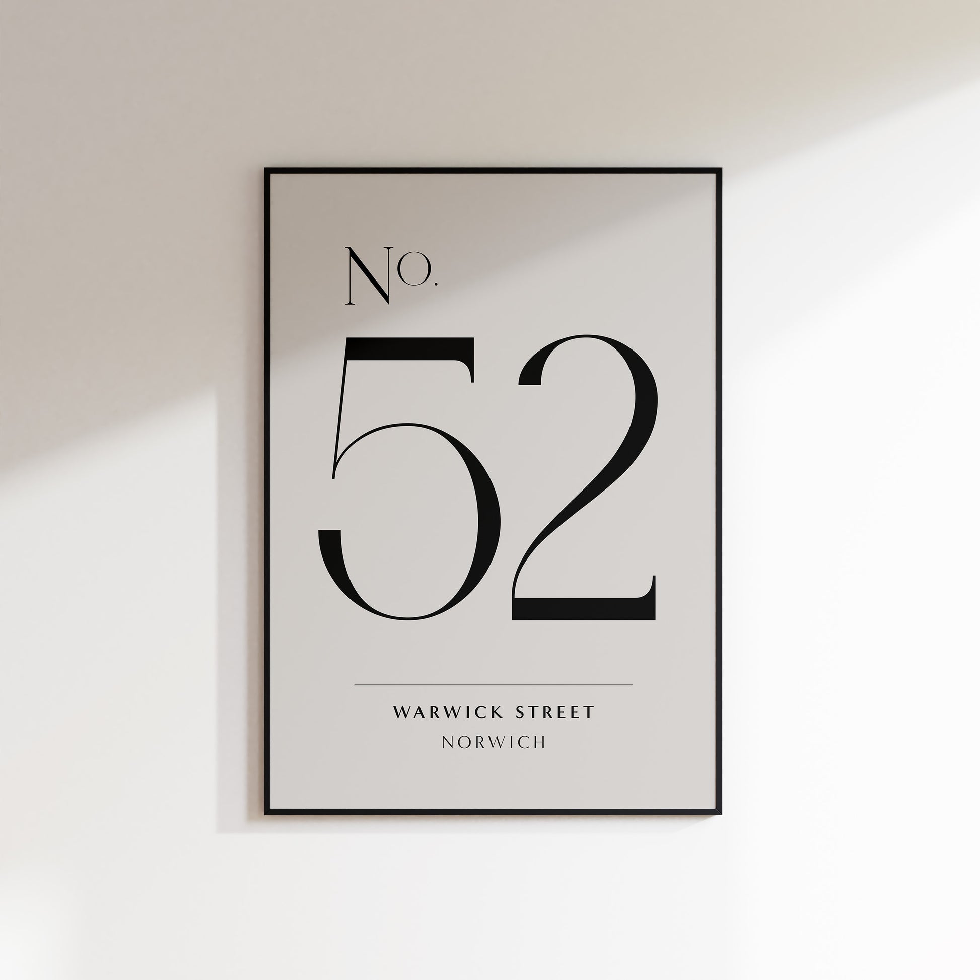 A house number print for the home, ideal for hallways, living rooms and bedrooms. This is a neutral beige coloured print with a slate coloured house number in a deco style serif typeface, and the street name and location underneath. Print is in a black frame on a white wall background.